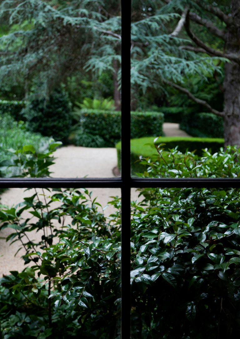 View of a landscaped garden path through a window with lush greenery and manicured hedges