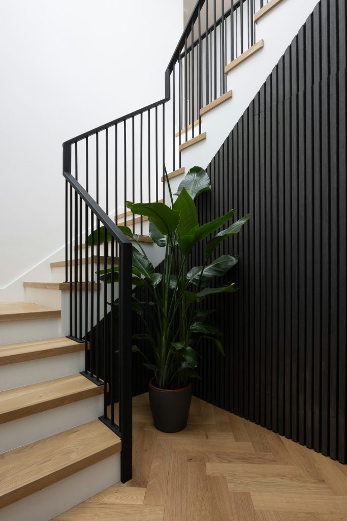 Contemporary staircase with oak treads, black metal balustrade, and feature wall panelling, complemented by a potted green plant.